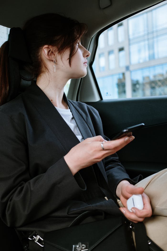 Home A woman with a ponytail and earrings uses a smartphone in the back of a car, focusing outside.