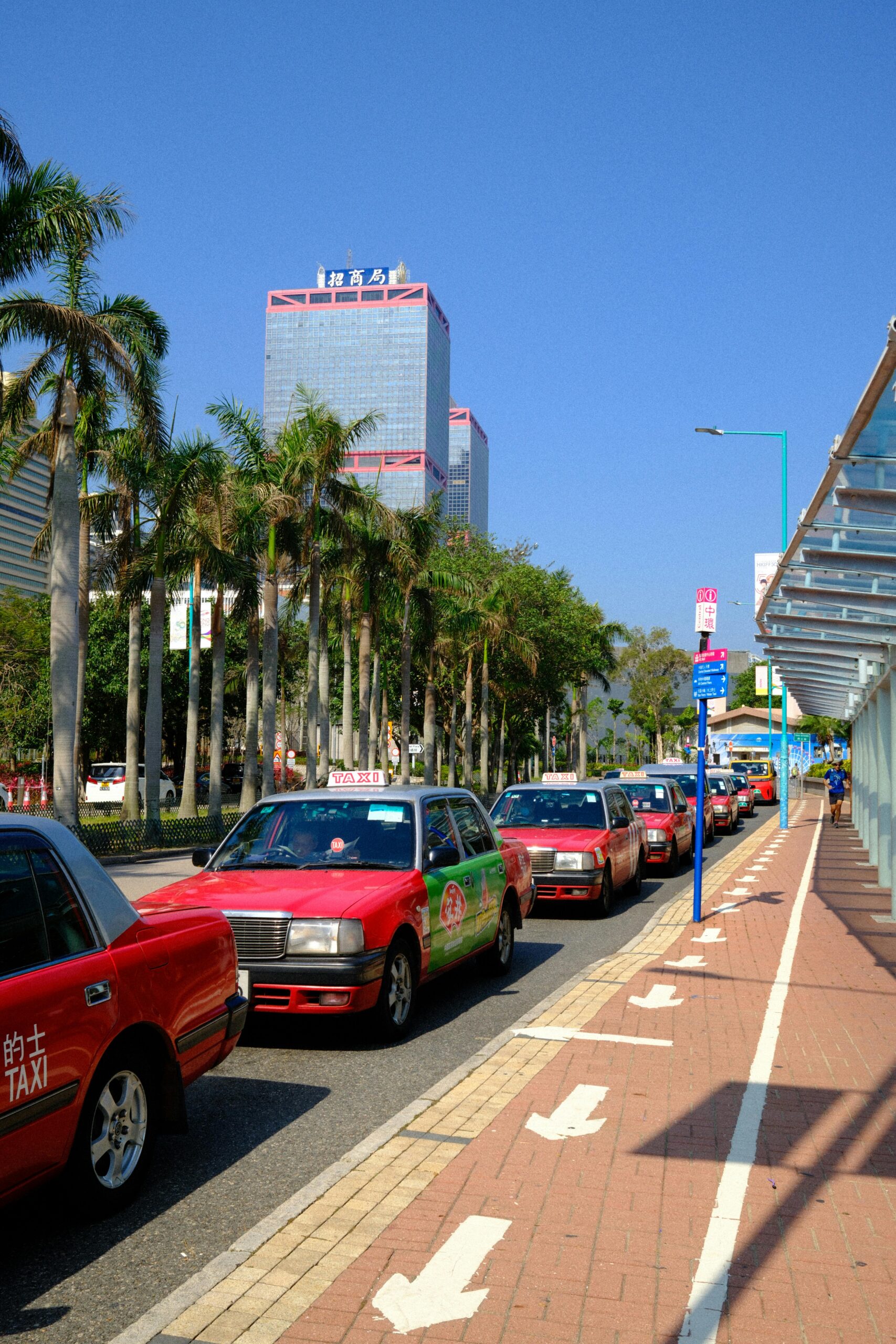 Red taxis line the streets of Hong Kong under sunny skies, with palm trees and skyscrapers in the background.