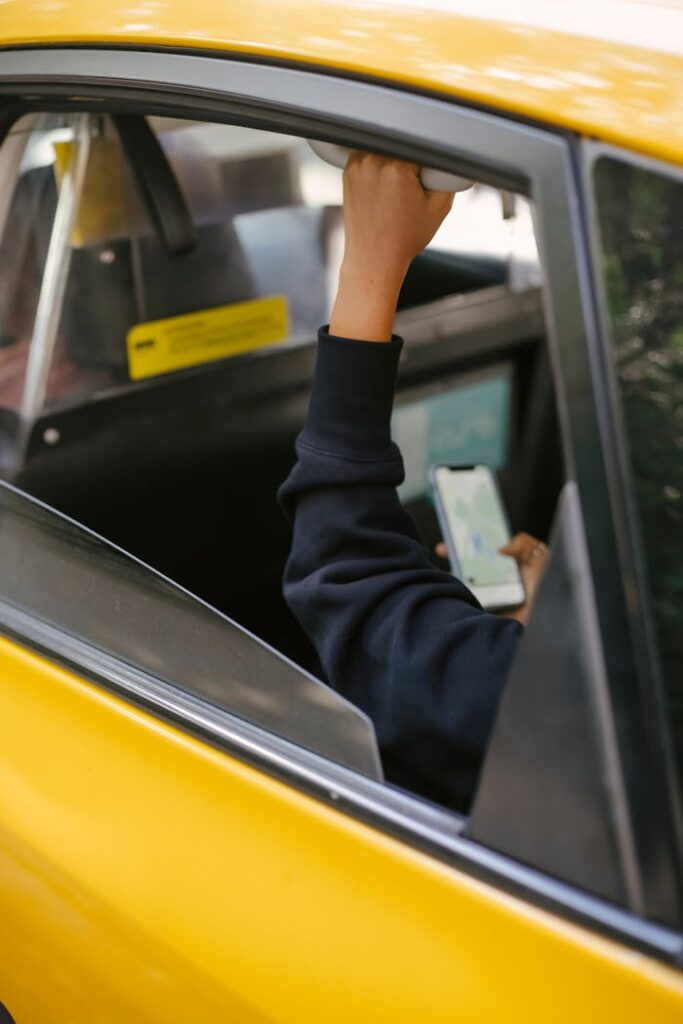 Unrecognizable passenger sitting on backseat and browsing smartphone while holding handle inside of yellow cab with lowered window during ride