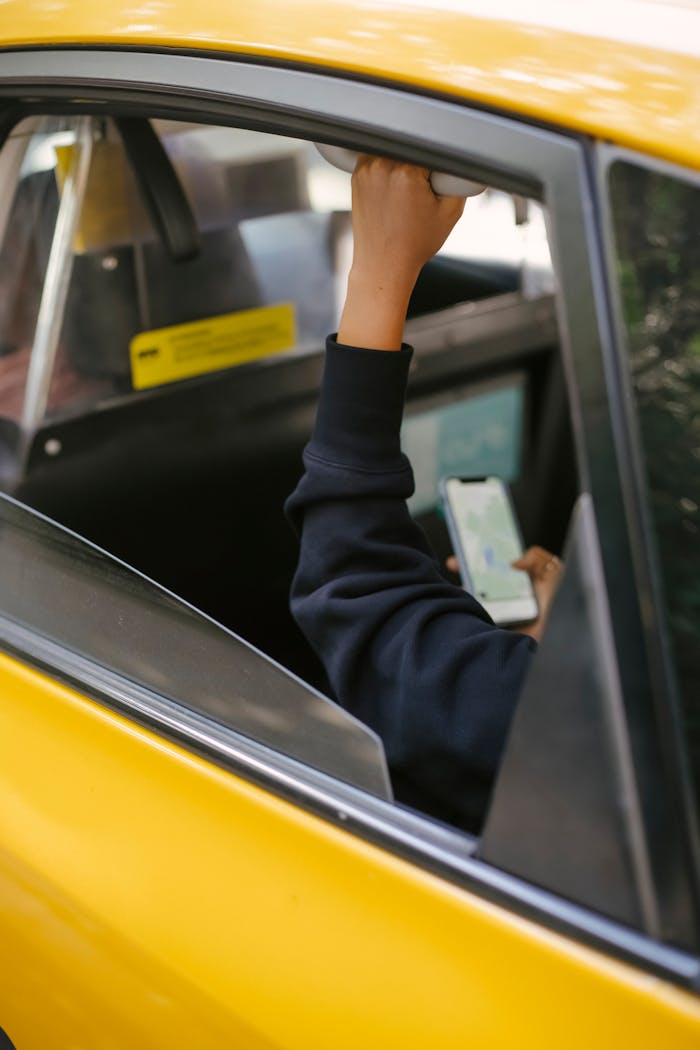Unrecognizable passenger sitting on backseat and browsing smartphone while holding handle inside of yellow cab with lowered window during ride